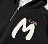 نمای نزدیک چاپ M Love روی هودی مشکی بچگانه و جزئیات زیپ و پارچه