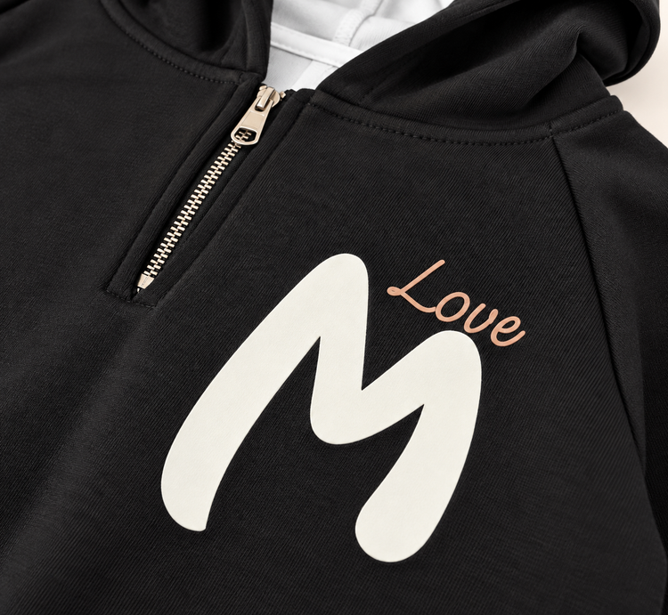 نمای نزدیک چاپ M Love روی هودی مشکی بچگانه و جزئیات زیپ و پارچه